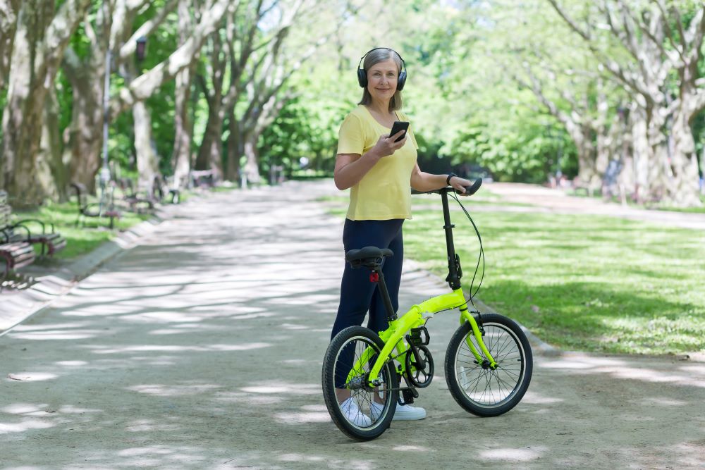 A woman holding a yellow bicycle while listening to her headphones and mobile device
